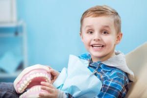 happy blonde boy in a dental chair holding a giant model of a jaw with teeth